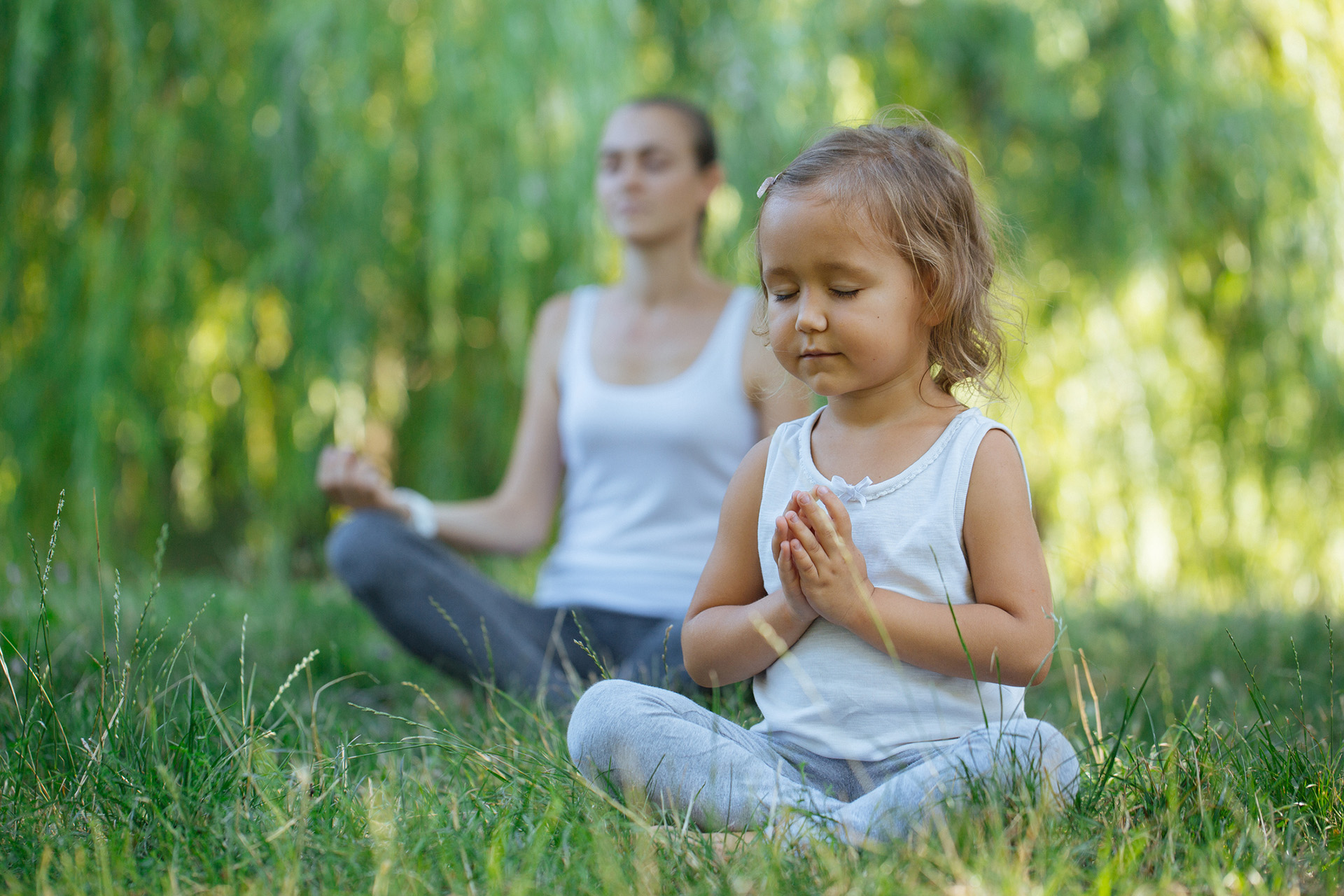 Young mother and cute little daughter meditating in lotus pose together at green summer park.